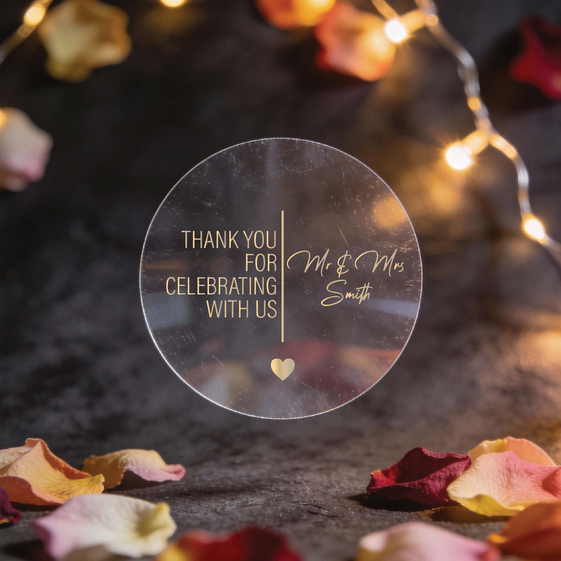 Circular clear sticker with gold foil text reading "Thank you for celebrating with us, Mr & Mrs Smith." Surrounded by rose petals and warm string lights.