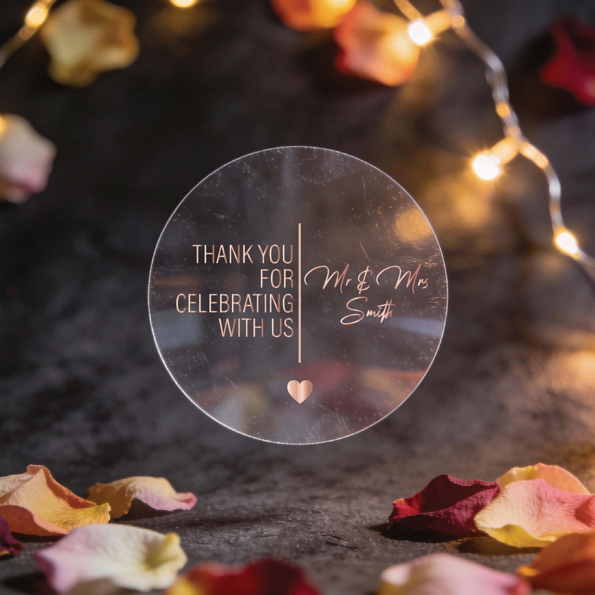 Circular clear sticker with rose gold foil text reading "Thank you for celebrating with us, Mr & Mrs Smith." Surrounded by rose petals and warm string lights.