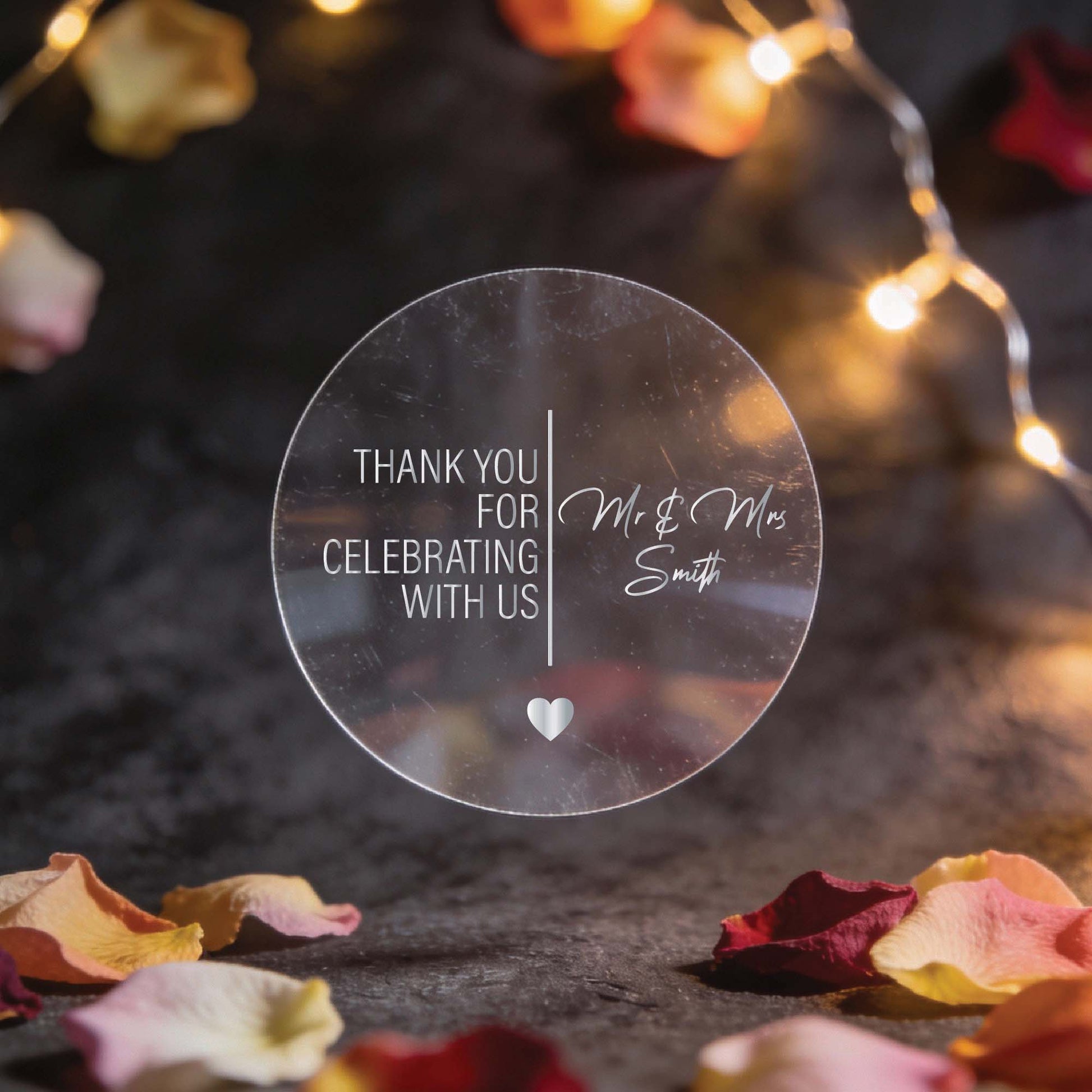 Circular clear sticker with silver foil text reading "Thank you for celebrating with us, Mr & Mrs Smith." Surrounded by rose petals and warm string lights.