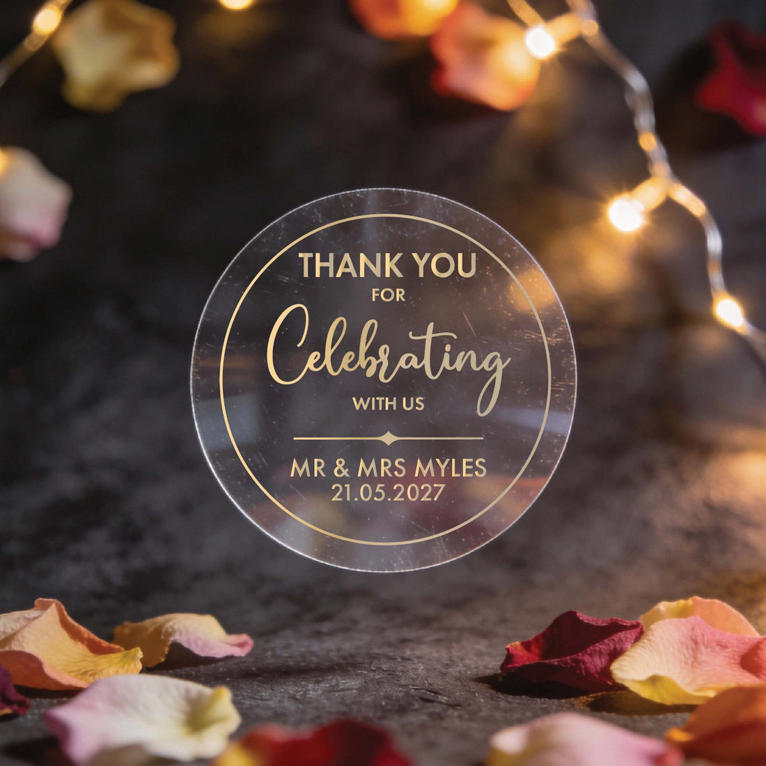 A hand holds a transparent circular token with gold foil text reading "Thank you for celebrating with us, Mr. & Mrs. Myles, 21.05.2027." Soft lights and flower petals in the blurred background create a warm, festive tone.