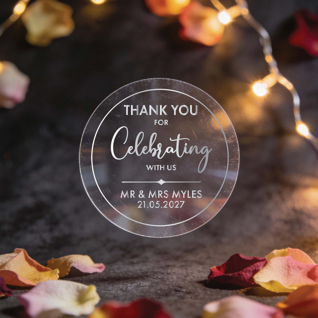 A hand holds a transparent circular token with gold foil text reading "Thank you for celebrating with us, Mr. & Mrs. Myles, 21.05.2027." Soft lights and flower petals in the blurred background create a warm, festive tone.