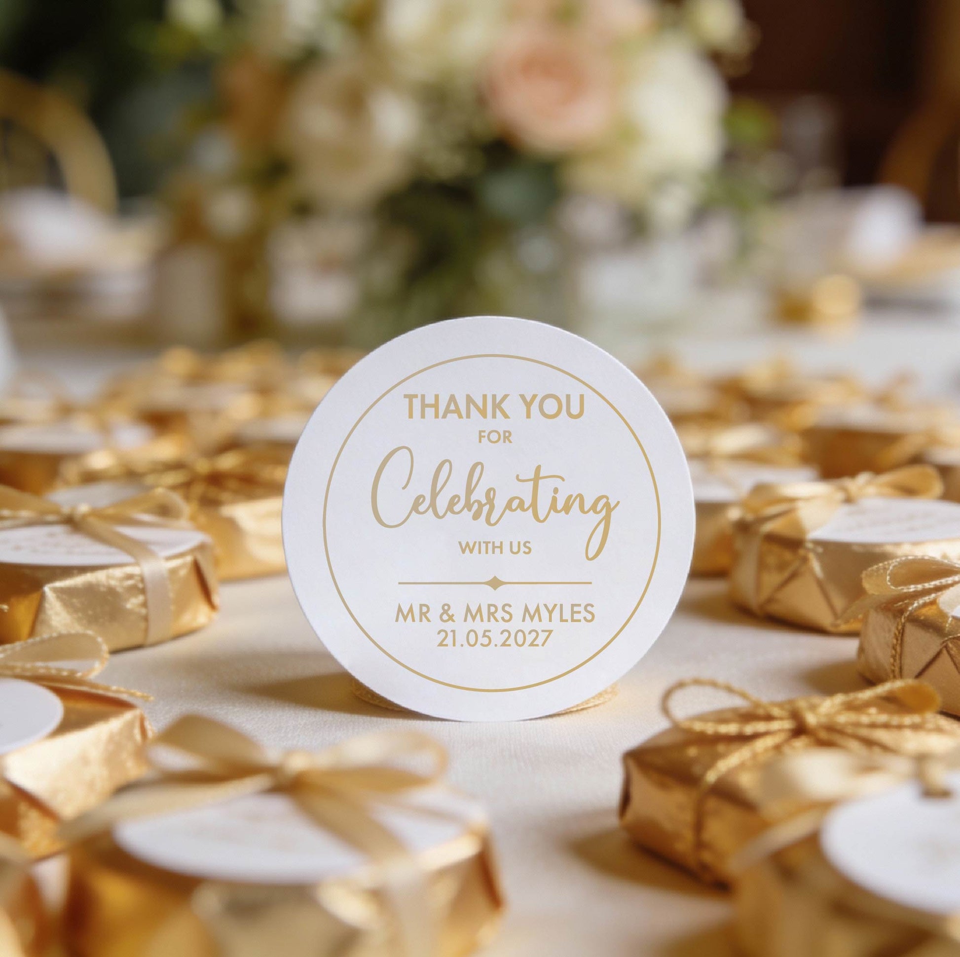 Round gloss sticker with gold foil text saying, "Thank you for celebrating with us, Mr & Mrs Myles, 21.05.2027." Surrounded by small gold-wrapped gifts, soft floral background.