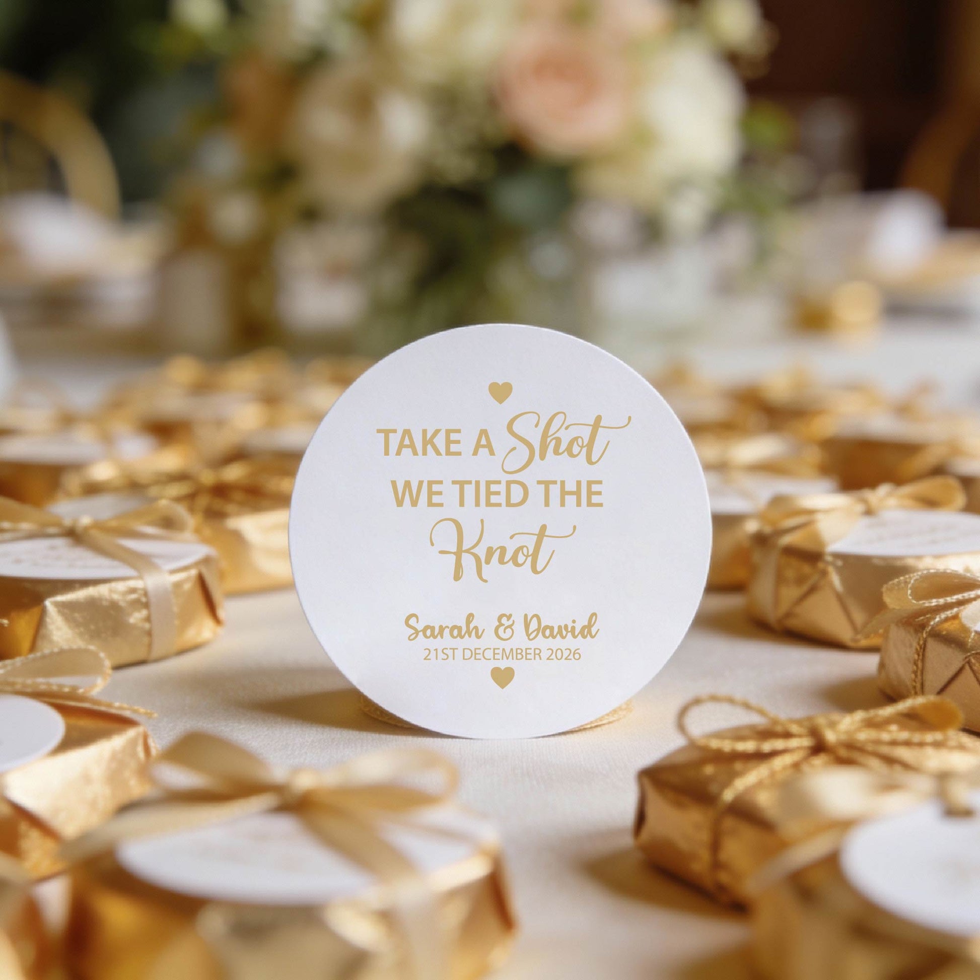 white gloss round sticker with gold foil text reads "Take a Shot We Tied the Knot", the names of the happy couple and the wedding date. The sticker is finished with two heart icons on the top and bottom of the sticker. 