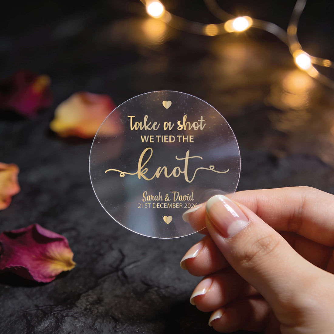 A transparent round sticker with gold foil text reading, "Take a shot, we tied the knot." The name of the happy couple and wedding date is written at the bottom with heart icons finishing the sticker at the top and bottom of the sticker. 