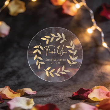 A round, clear sticker with gold foil leaf design and the words "Thank You," "Sarah & James," and "21 10 25." Surrounded by fairy lights and rose petals. Romantic ambiance.
