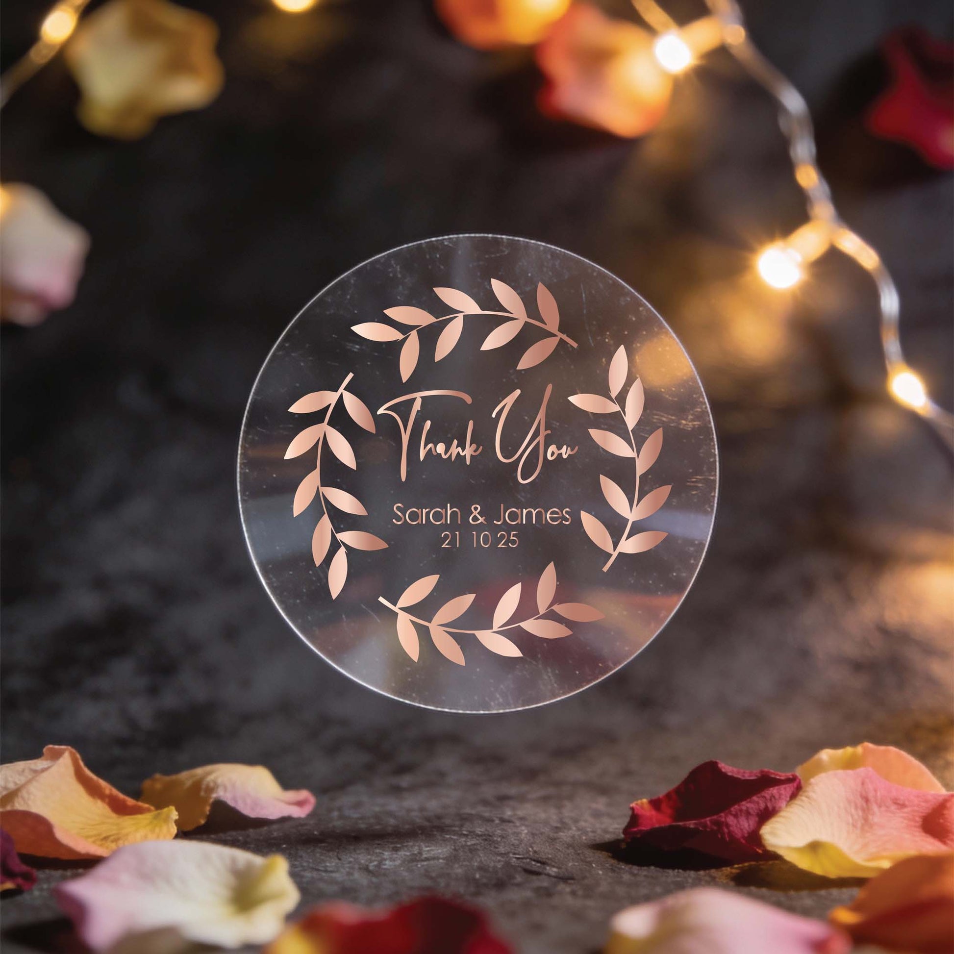 A round, clear sticker with rose gold foil leaf design and the words "Thank You," "Sarah & James," and "21 10 25." Surrounded by fairy lights and rose petals. Romantic ambiance.