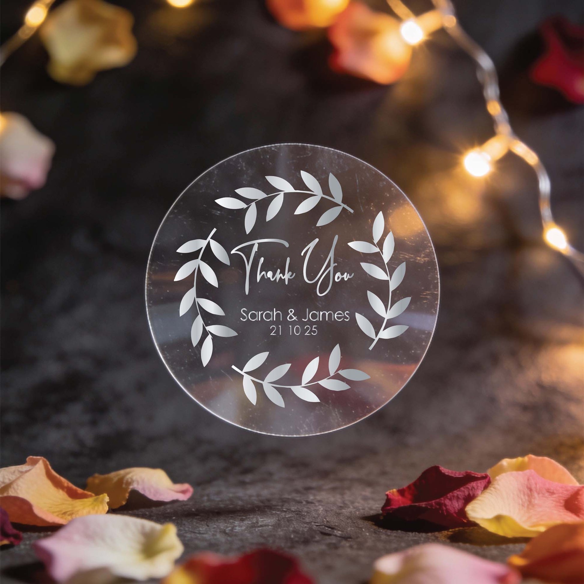 A round, clear sticker with silver foil leaf design and the words "Thank You," "Sarah & James," and "21 10 25." Surrounded by fairy lights and rose petals. Romantic ambiance.