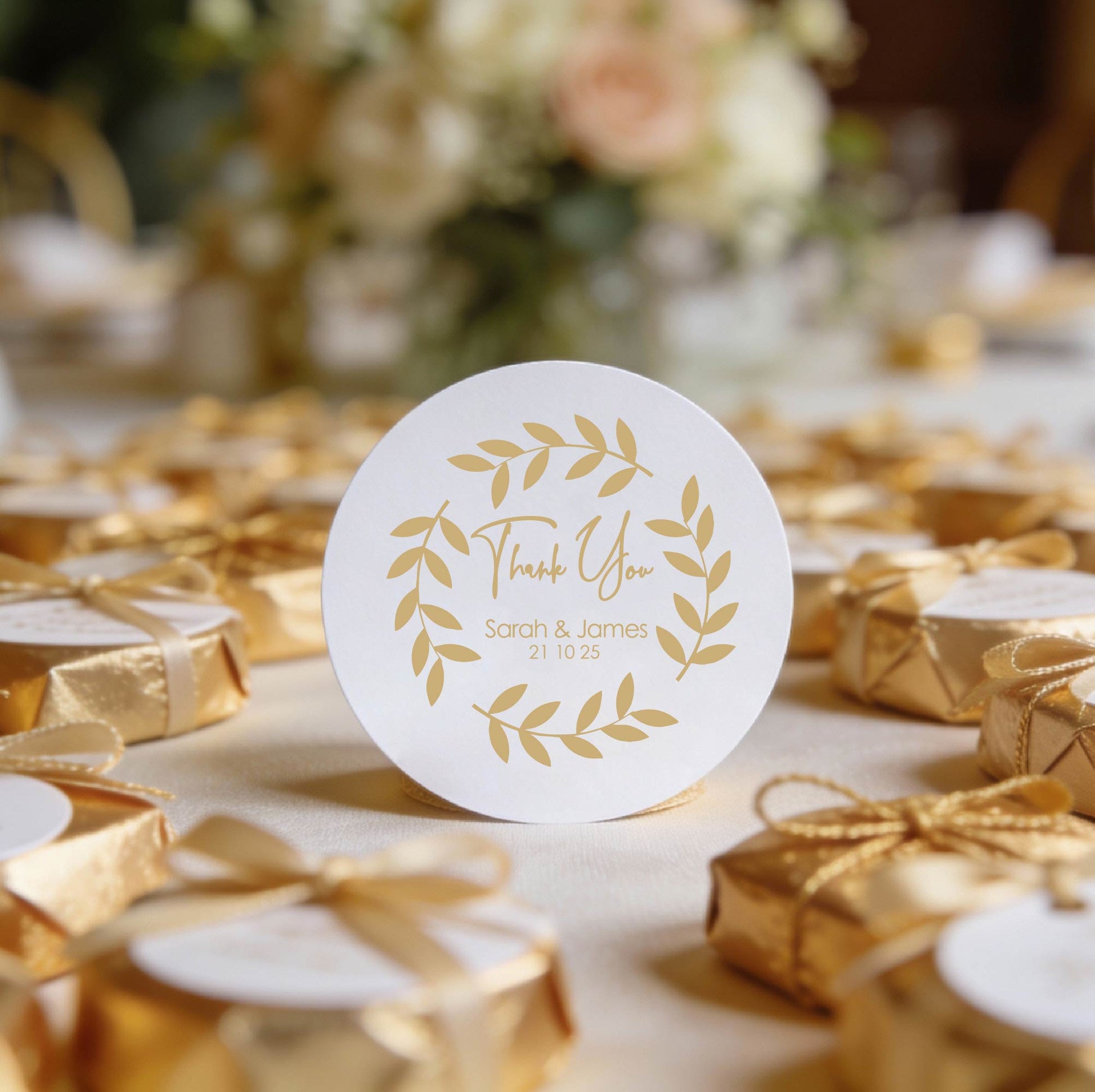 A round, white gloss sticker with gold foil leaf design and the words "Thank You," "Sarah & James," and "21 10 25." Surrounded by fairy lights and rose petals. Romantic ambiance.