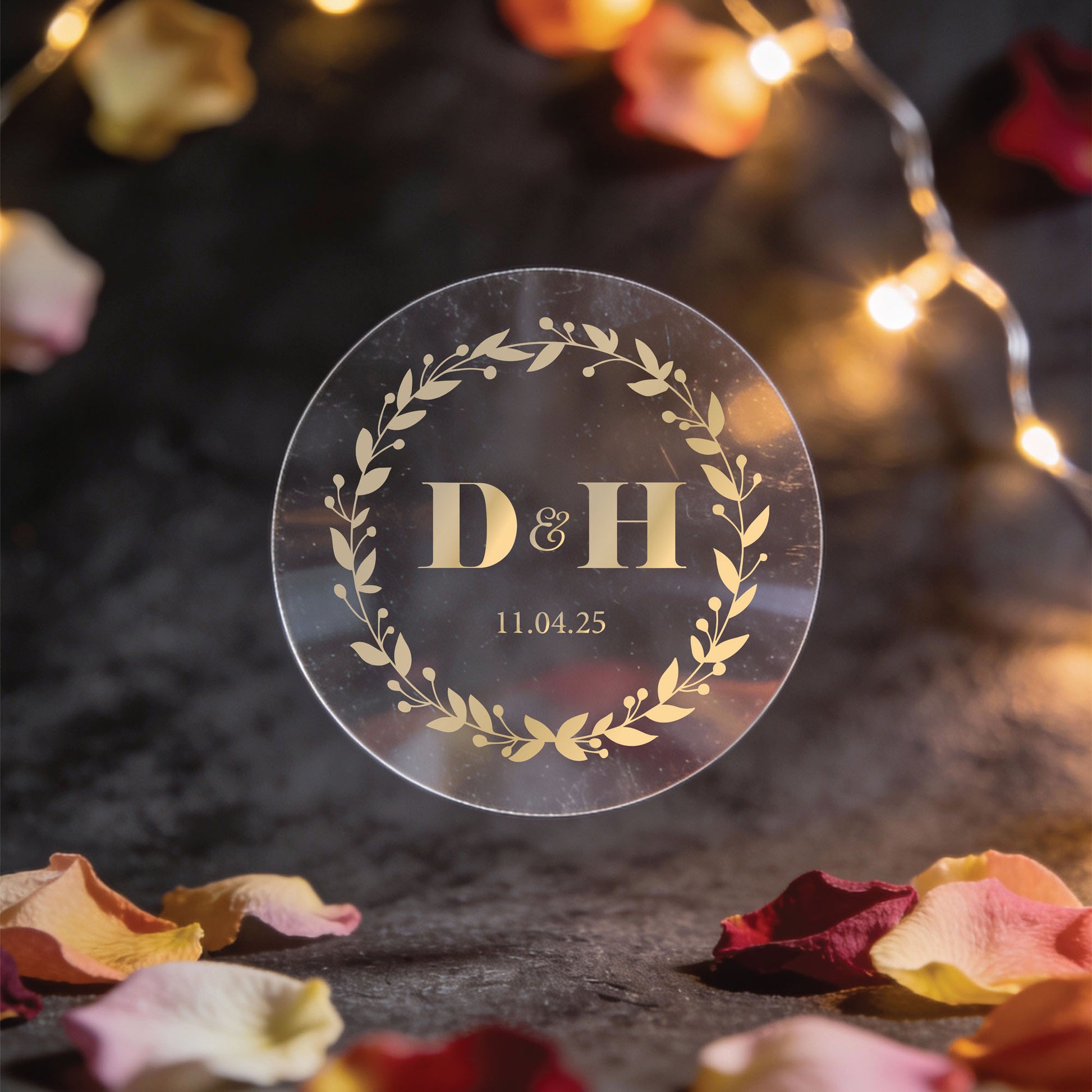 A round Transparent sticker with gold  foil print with initials "D & H" and date "11.04.25" encircled by a laurel wreath, set against warm string lights and rose petals.