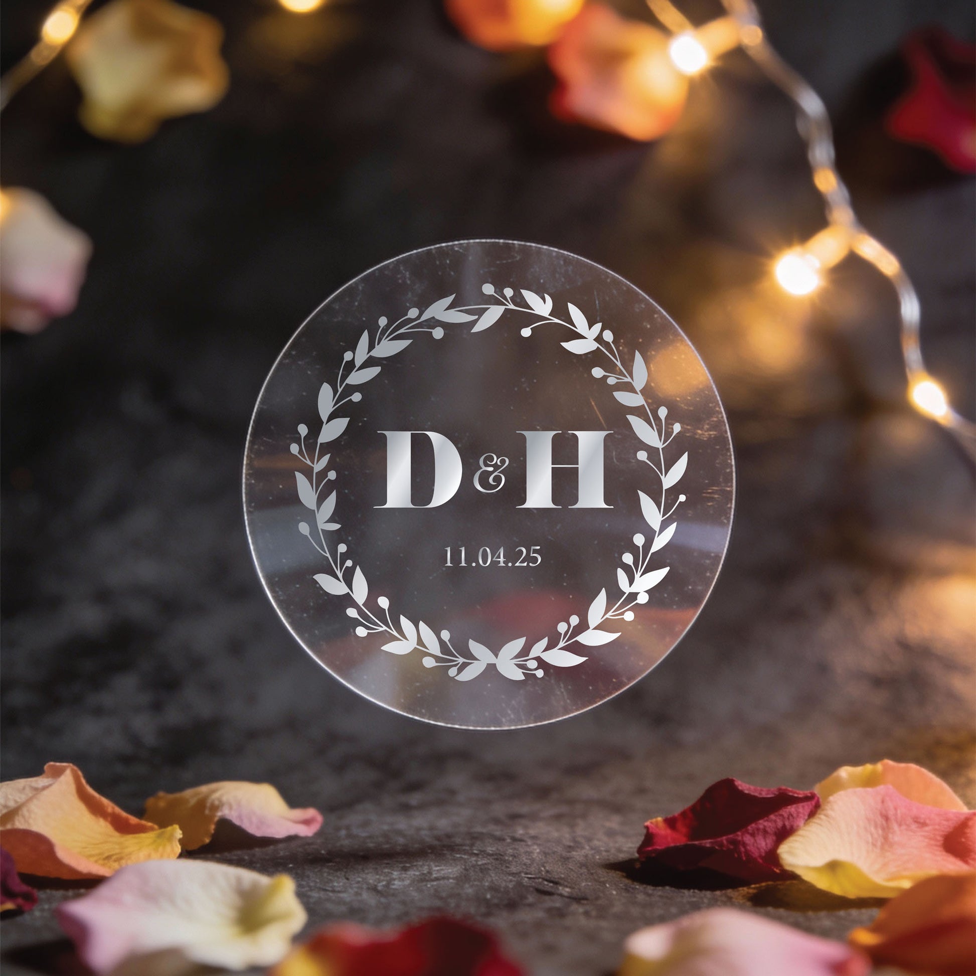 A round Transparent sticker with silver foil print with initials "D & H" and date "11.04.25" encircled by a laurel wreath, set against warm string lights and rose petals.