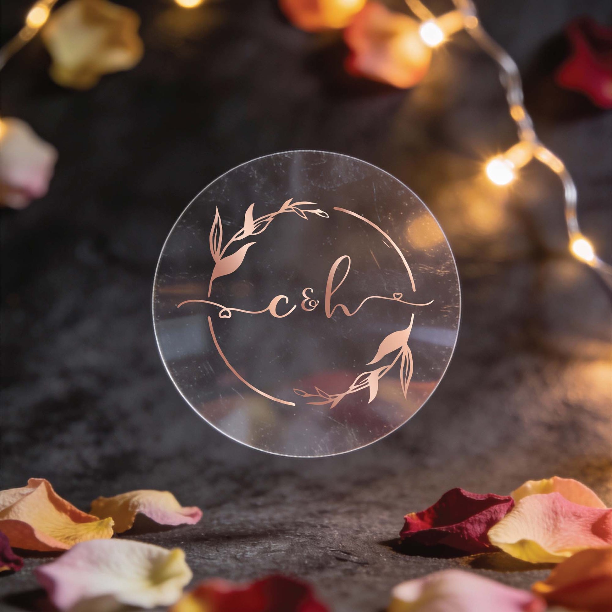 A round, transparent sticker with the initials "c&h" in elegant rose gold foil lettering, surrounded by decorative leaves. Warm fairy lights and scattered rose petals create a romantic ambiance.