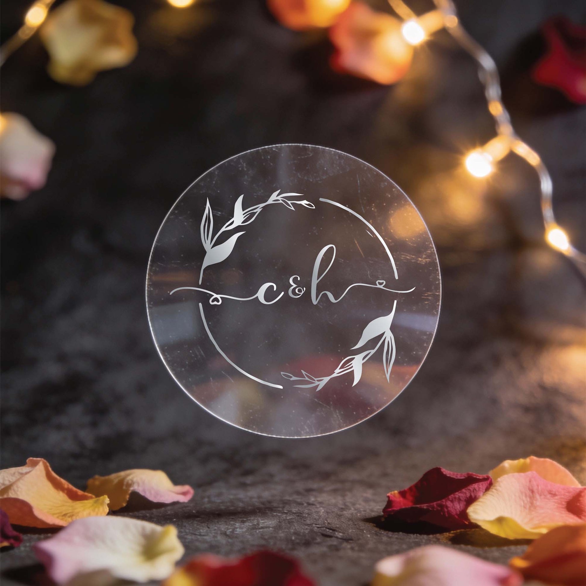 A round, transparent sticker with the initials "c&h" in elegant silver foil lettering, surrounded by decorative leaves. Warm fairy lights and scattered rose petals create a romantic ambiance.