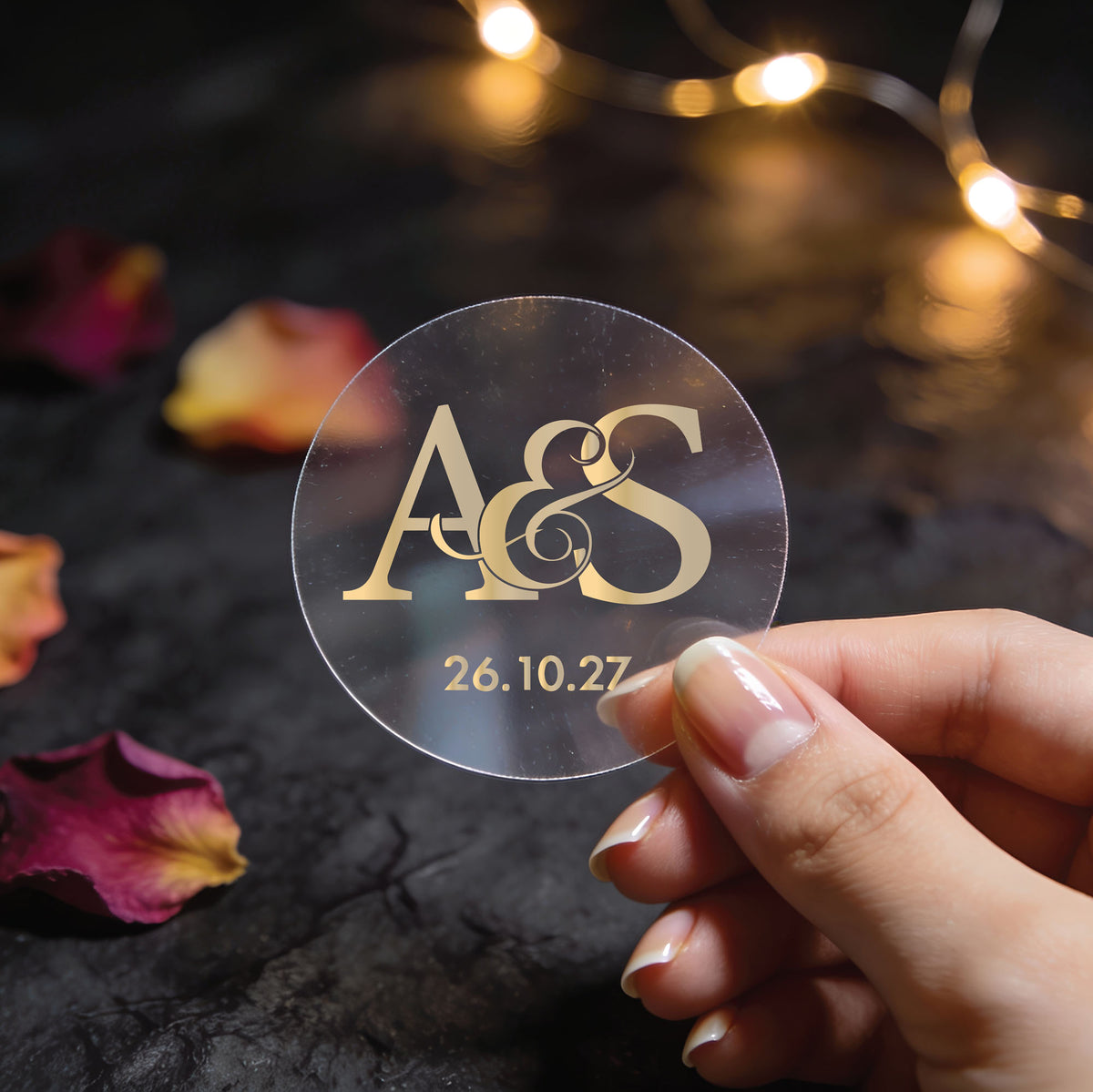 Clear transparent gold foil monogram wedding sticker with large ampersand, entwined initials and wedding date, held above scattered rose petals and fairy lights.