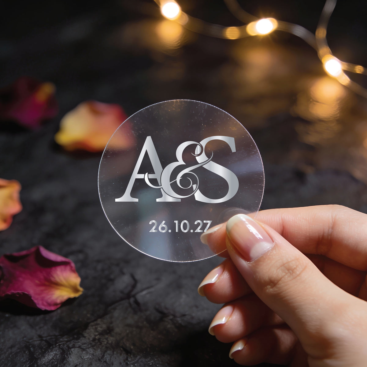 Clear transparent silver foil monogram wedding sticker with large ampersand, entwined initials and wedding date, held above scattered rose petals and fairy lights.