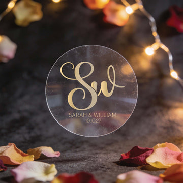 A transparent round sticker with "SW" in elegant gold foil script and "Sarah & William 10.10.27" below. Warm lights and rose petals create a romantic setting.