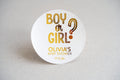 A round white sticker with gold foil text asking 'Boy or Girl?' followed by 'OLIVIA'S BABY SHOWER' and a date. The sticker is placed on a neutral background.