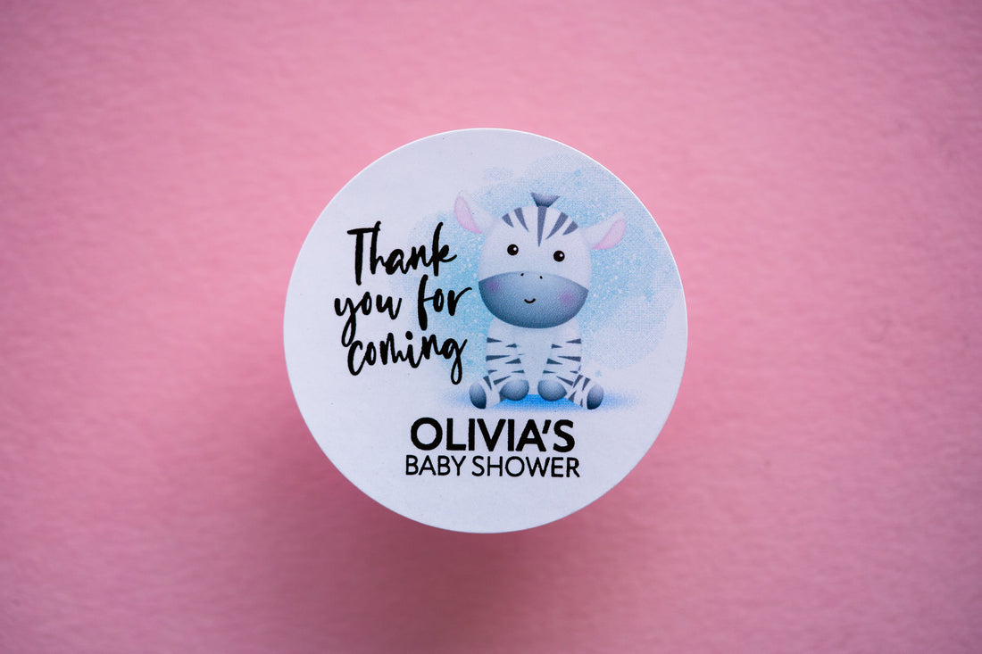 A baby shower sticker with a lovely colour print of an zebra sitting on the floor. Thank you for coming is written in a handwriting font next to the zebra and the mother to be name on the bottom. The writing is finished with metallic foil