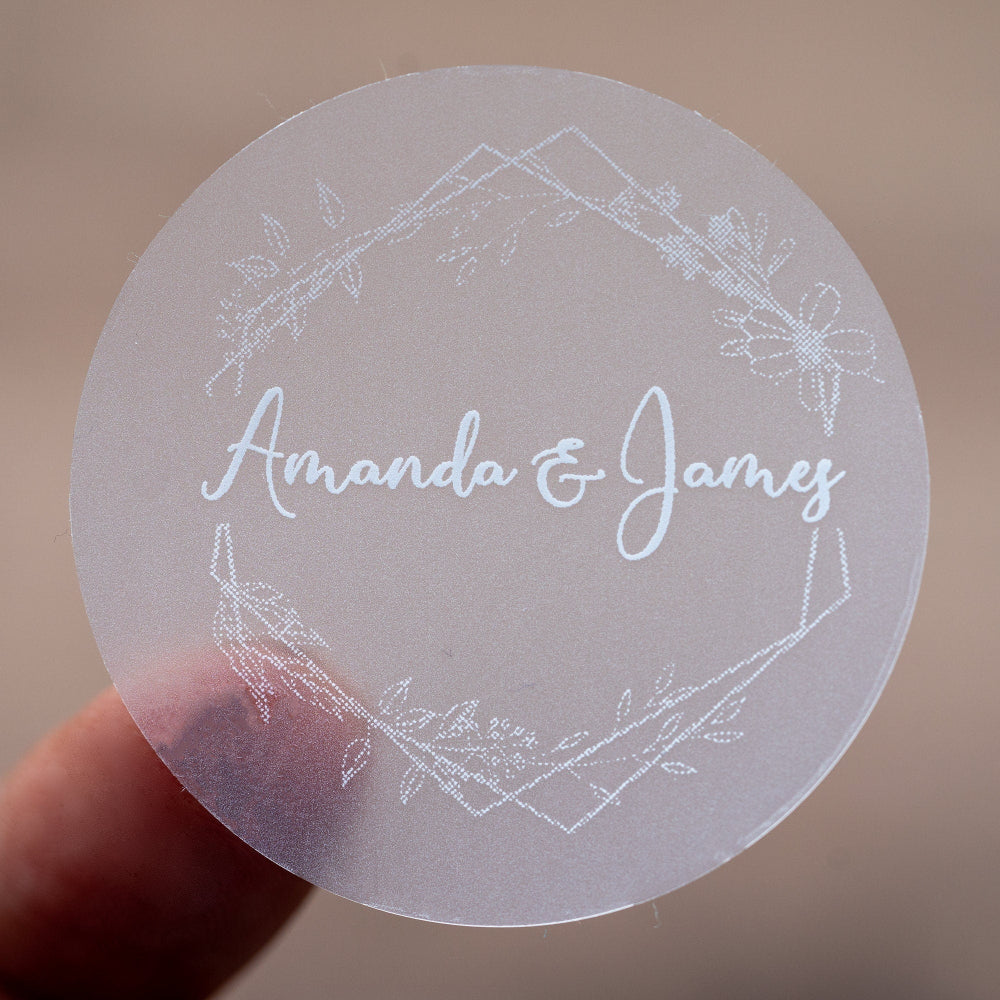 A round, white-print custom sticker with the names 'Amanda & James' printed in the centre surrounded by a floral leaf frame design.