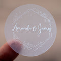 A round, white-print custom sticker with the names 'Amanda & James' printed in the centre surrounded by a floral leaf frame design.