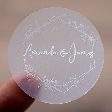 A round, white-print custom sticker with the names 'Amanda & James' printed in the centre surrounded by a floral leaf frame design.