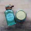 A round, white-print custom sticker with the names 'Amanda & James' printed in the centre surrounded by a floral leaf frame design on a Blue bottle and jar with 'Amanda & Jenny'.