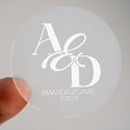A clear round sticker with the monogram 'A & D' and the names 'Amanda & David' printed in white ink, held between fingers.