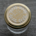 Gold lid with a custom round white sticker with a 'Thank You' message, leaf design, and personalised text including names and a date.