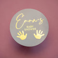 A round grey sticker with the text 'Baby Shower' and a pair of hand prints in gold foil. The name 'Emma' is written in a cursive script at the top.