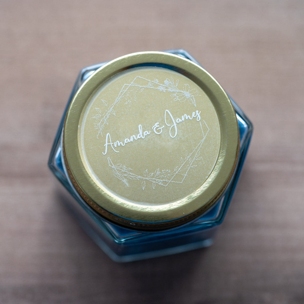 A round, white-print custom sticker with the names 'Amanda & James' printed in the centre surrounded by a floral leaf frame design.
