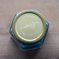 A round, white-print custom sticker with the names 'Amanda & James' printed in the centre surrounded by a floral leaf frame design on a gold lid of a candle.