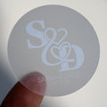 A close-up of a round clear sticker with a custom white monogram print of a couple's initials and names.