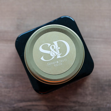 A round, white monogram sticker with a gold border applied to a clear glass jar lid.