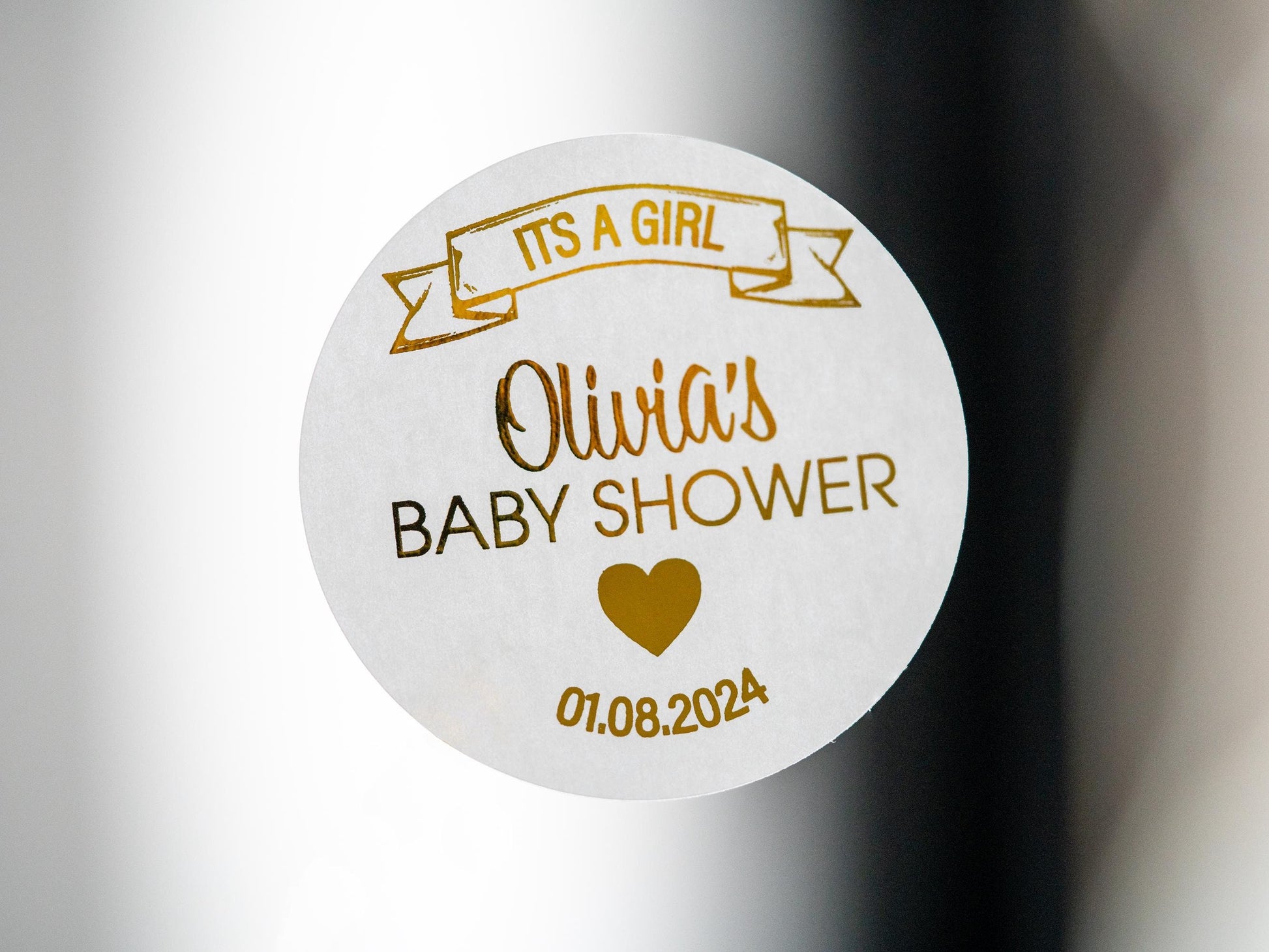 The name of the mummy-to-be in the centre of the sticker using a lovely hand written font. A banner announcing the sex of the baby on the top of the sticker and the date of the baby shower on the bottom. The whole design embossed in metallic foil