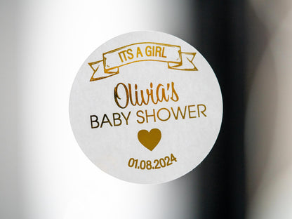 The name of the mummy-to-be in the centre of the sticker using a lovely hand written font. A banner announcing the sex of the baby on the top of the sticker and the date of the baby shower on the bottom. The whole design embossed in metallic foil
