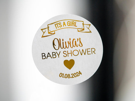 The name of the mummy-to-be in the centre of the sticker using a lovely hand written font. A banner announcing the sex of the baby on the top of the sticker and the date of the baby shower on the bottom. The whole design embossed in metallic foil