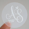 A close-up of a round, clear sticker with a white monogram print and personalized text 'Amanda & Sam'.