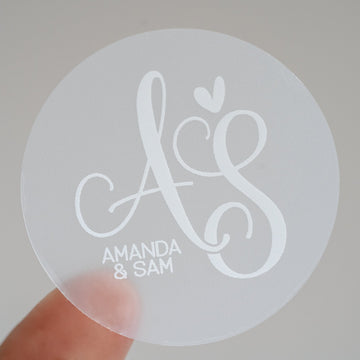 A close-up of a round, clear sticker with a white monogram print and personalized text 'Amanda & Sam'.