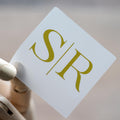 A square clear sticker with a monogram design in foil gold, placed on a textured background.