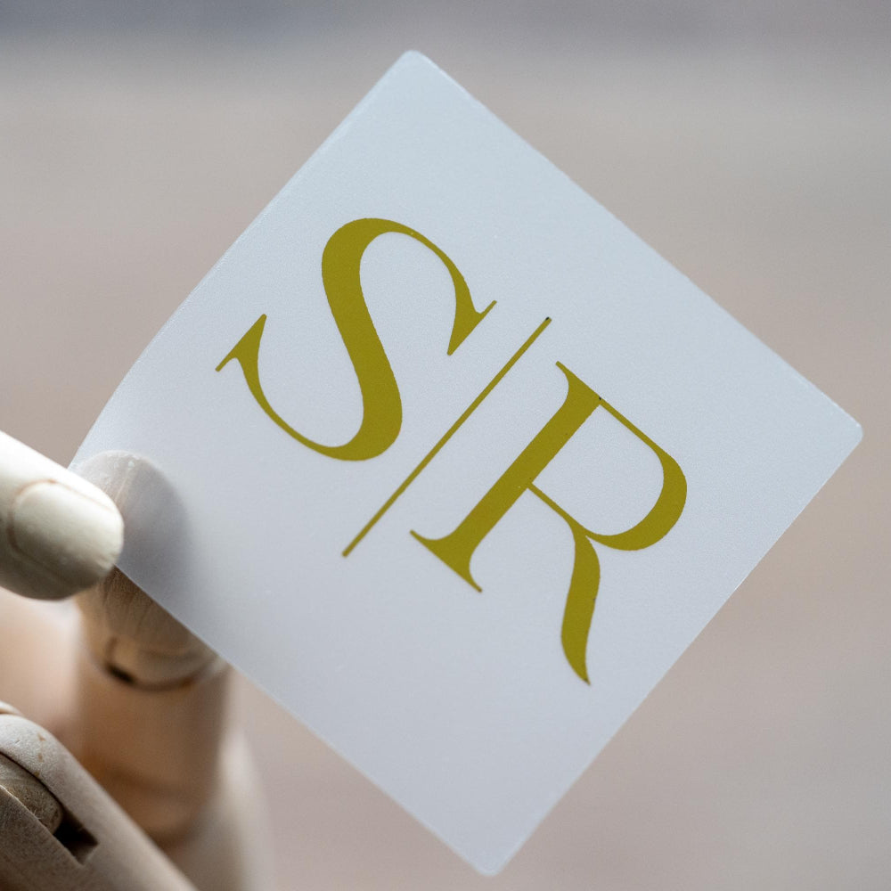A square clear sticker with a monogram design in foil gold, placed on a textured background.