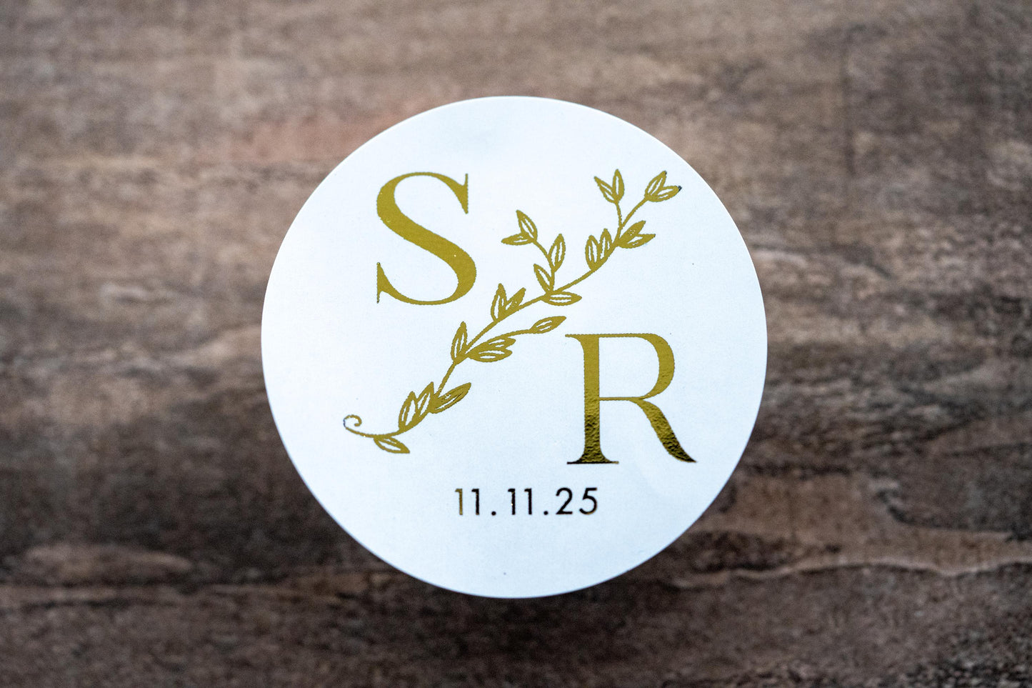 A custom round monogram sticker with a leaf border line running diagonally across the design. The initials of the bride and groom sit either side of the leaf border line with the date at the bottom. The design is finished with a lovely metallic foil.