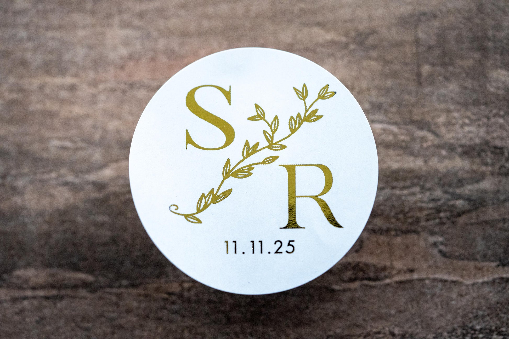 A custom round monogram sticker with a leaf border line running diagonally across the design. The initials of the bride and groom sit either side of the leaf border line with the date at the bottom. The design is finished with a lovely metallic foil.