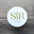 A custom white gloss round monogram sticker with a leaf border line running vertically across the middle. The initials of the bride and groom sit either side of the leaf border line with the date at the bottom. The design is finished with a lovely metallic foil.