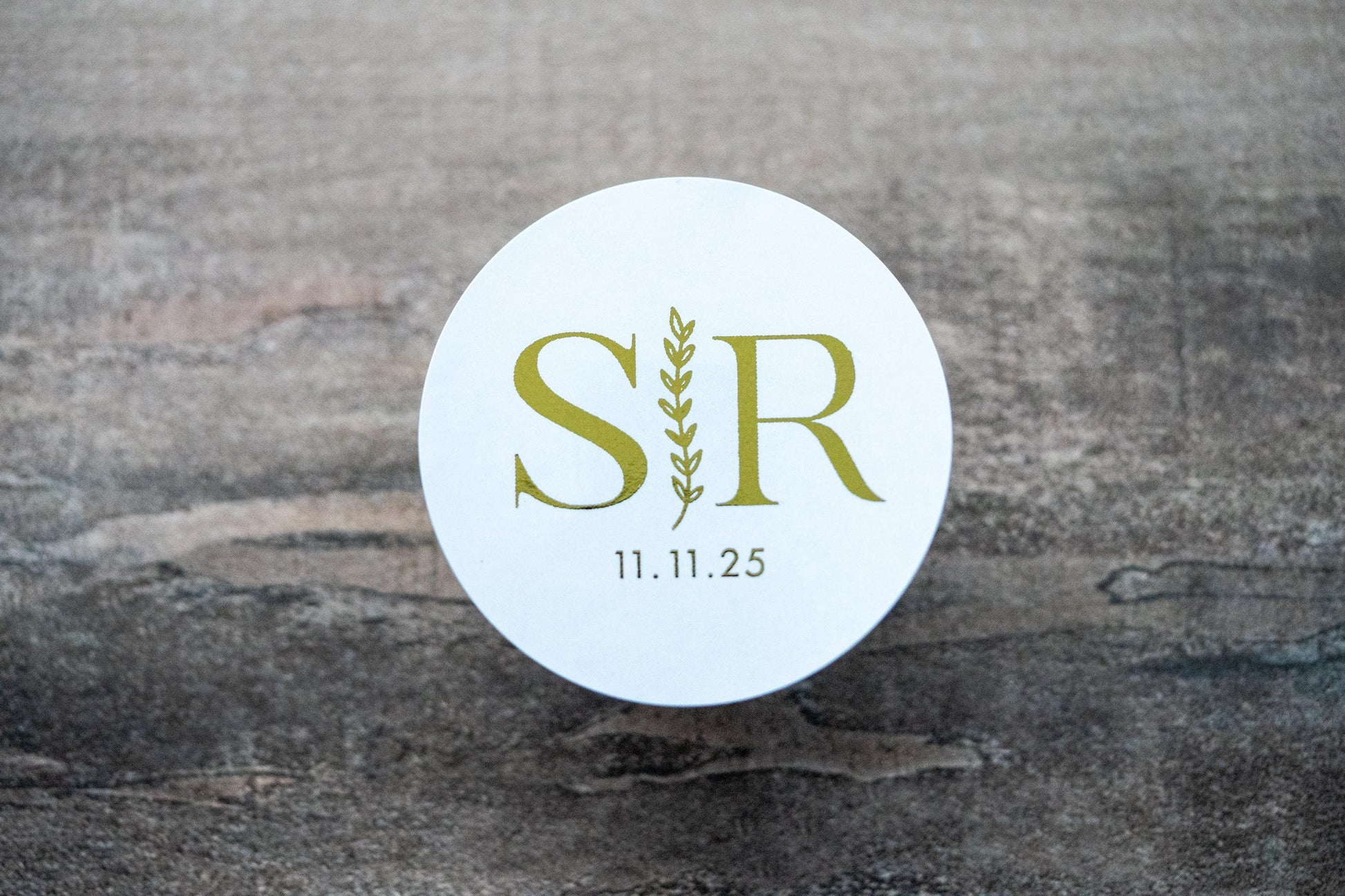 A custom round monogram sticker with a leaf border line running vertically across the middle. The initials of the bride and groom sit either side of the leaf border line with the date at the bottom. The design is finished with a lovely metallic foil.