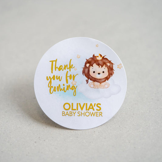 A baby shower sticker with a lovely colour print of a lion sitting on clouds. Thank you for coming is written in a handwriting font next to the lion and the mother to be name on the bottom. The writing is finished with metallic foil