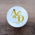 A round, white sticker with a gold foil monogram print of two letters, and the names 'Amanda & David' printed below, along with a date '10.10.24
