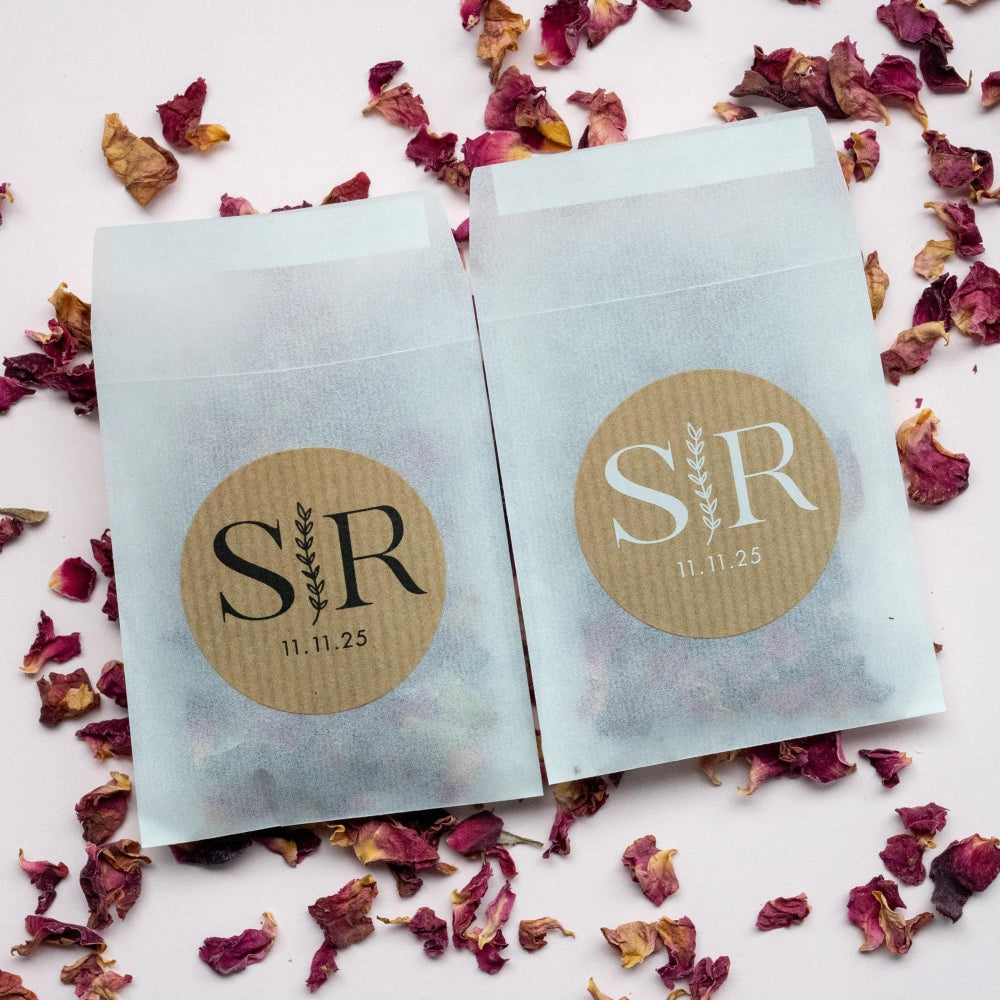 Custom kraft monogram wedding stickers with couple's initials and a leaf branch design, displayed on a table scattered with rose petals.