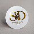 A close-up image of a round, white sticker with a gold foil monogram print of the names Sarah and David, along with a date.