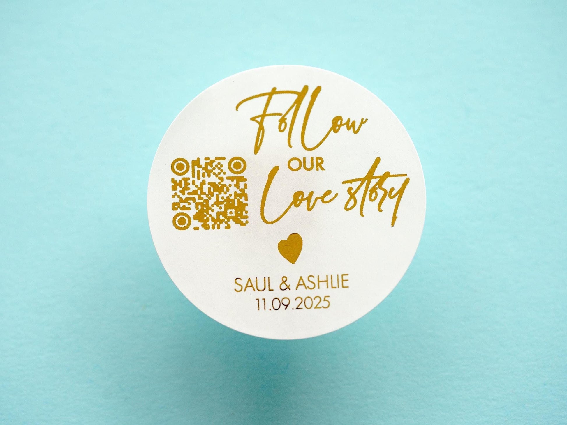 Save the date wedding sticker with a customisable QR code. Follow our love story is written in a handwritten font, a heart icon separates this and the name of the couple and date. The sticker is finished off with metallic foil for an added shine.