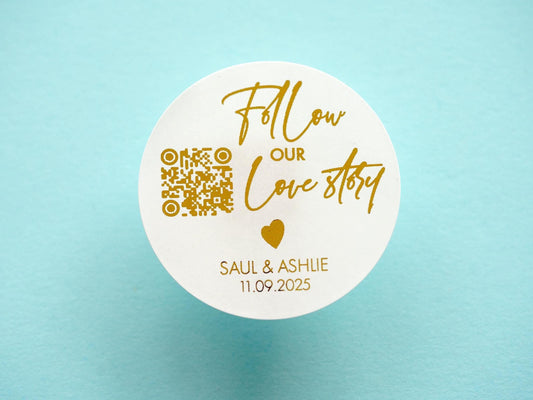Save the date wedding sticker with a customisable QR code. Follow our love story is written in a handwritten font, a heart icon separates this and the name of the couple and date. The sticker is finished off with metallic foil for an added shine.