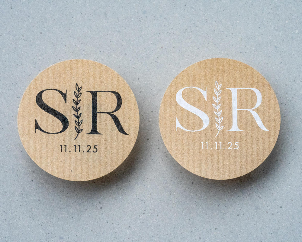 Custom kraft monogram wedding stickers with couple's initials and a leaf branch design, displayed on a table scattered with rose petals.